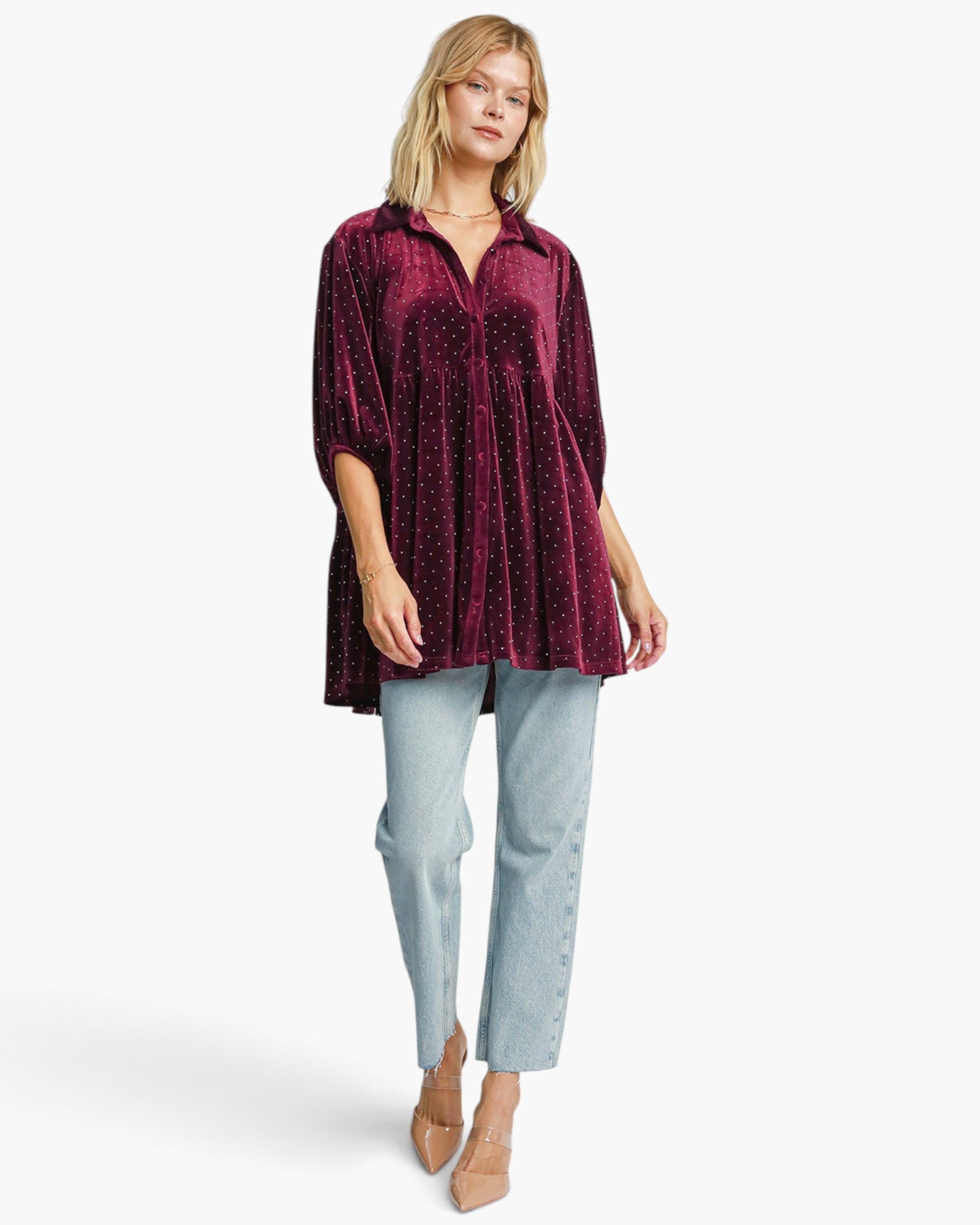 A woman modeling a deep burgundy velvet tunic top with sequined detailing, paired with light-wash blue jeans and beige heels, against a plain white studio background.