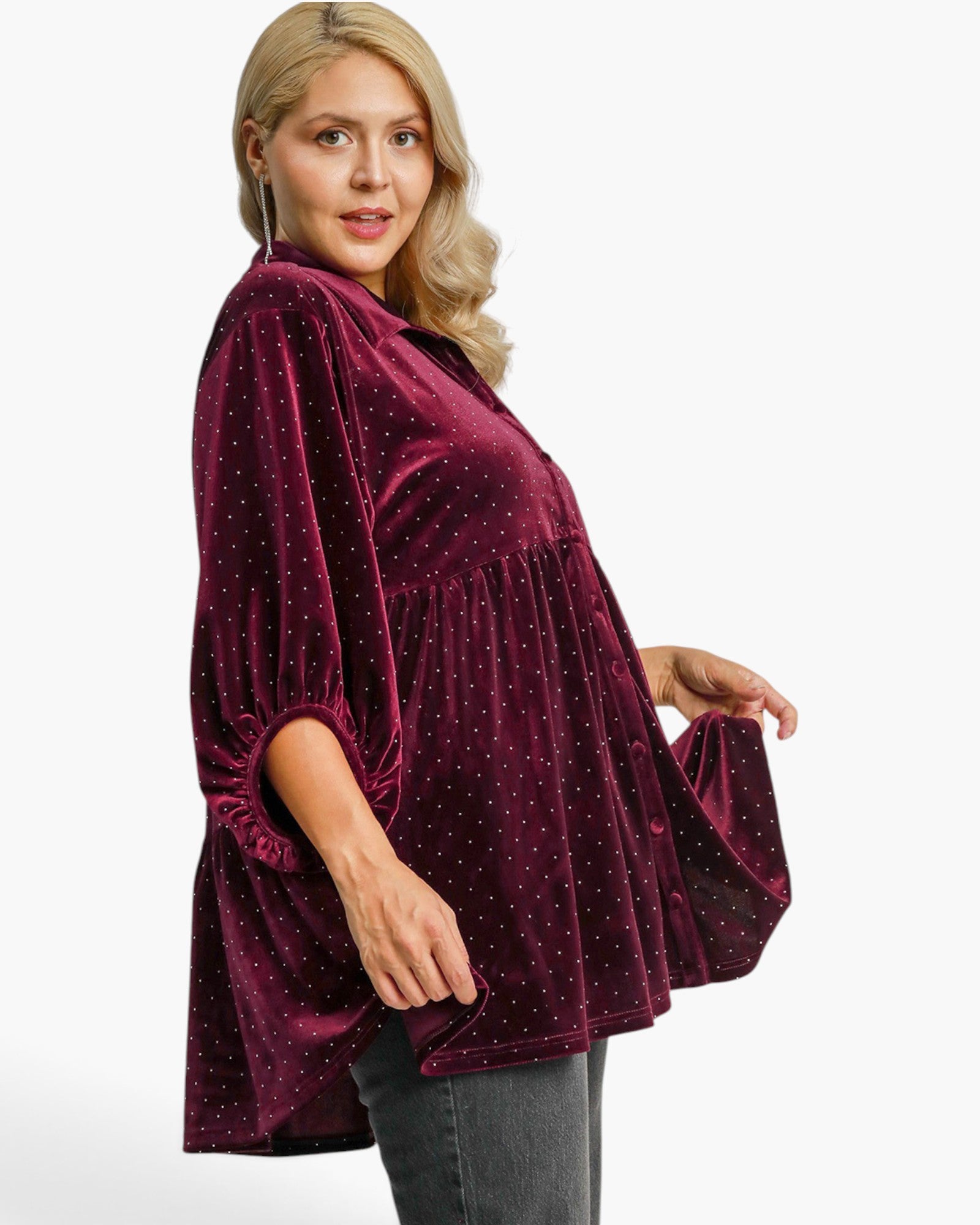 A woman wearing a burgundy velvet top with small gold polka dots, featuring a high collar, three-quarter sleeves, and a gathered peplum waist, paired with gray jeans against a plain white background.