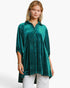 A woman modeling a teal green velvet tunic top with a subtle polka dot pattern, paired with white pants, against a plain white studio background.