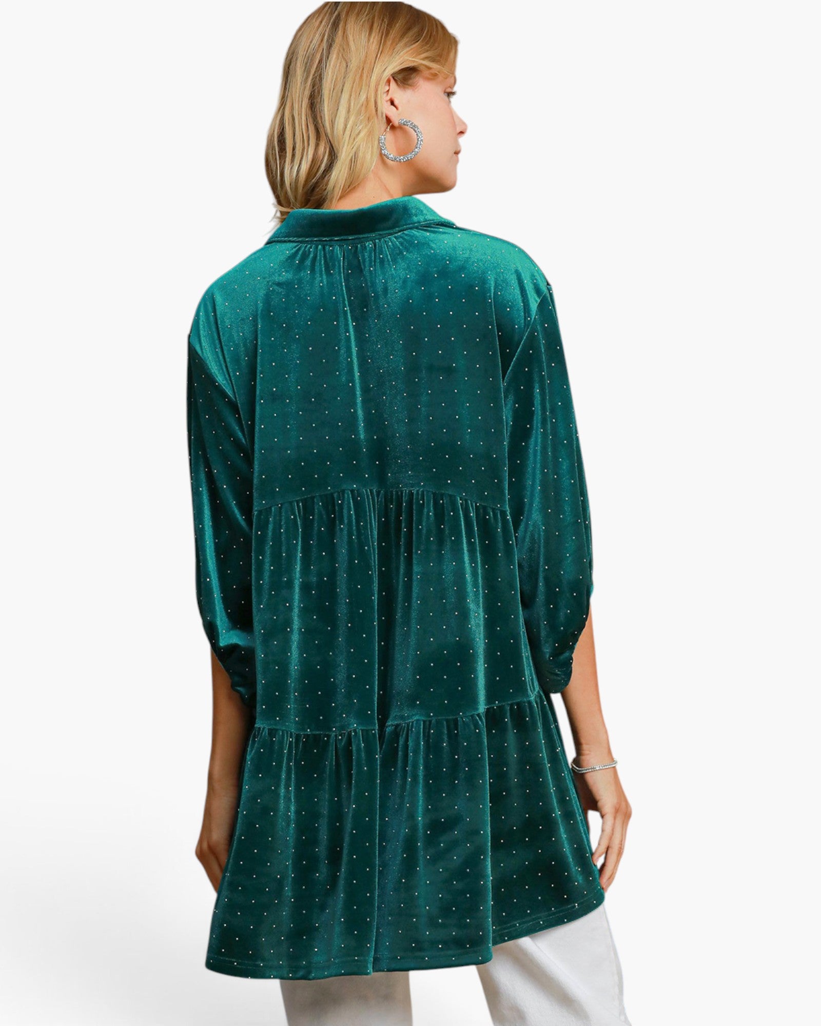 A woman shown from the back wearing a teal green velvet tiered tunic top with a subtle white polka dot pattern, paired with white pants, against a plain white studio background.
