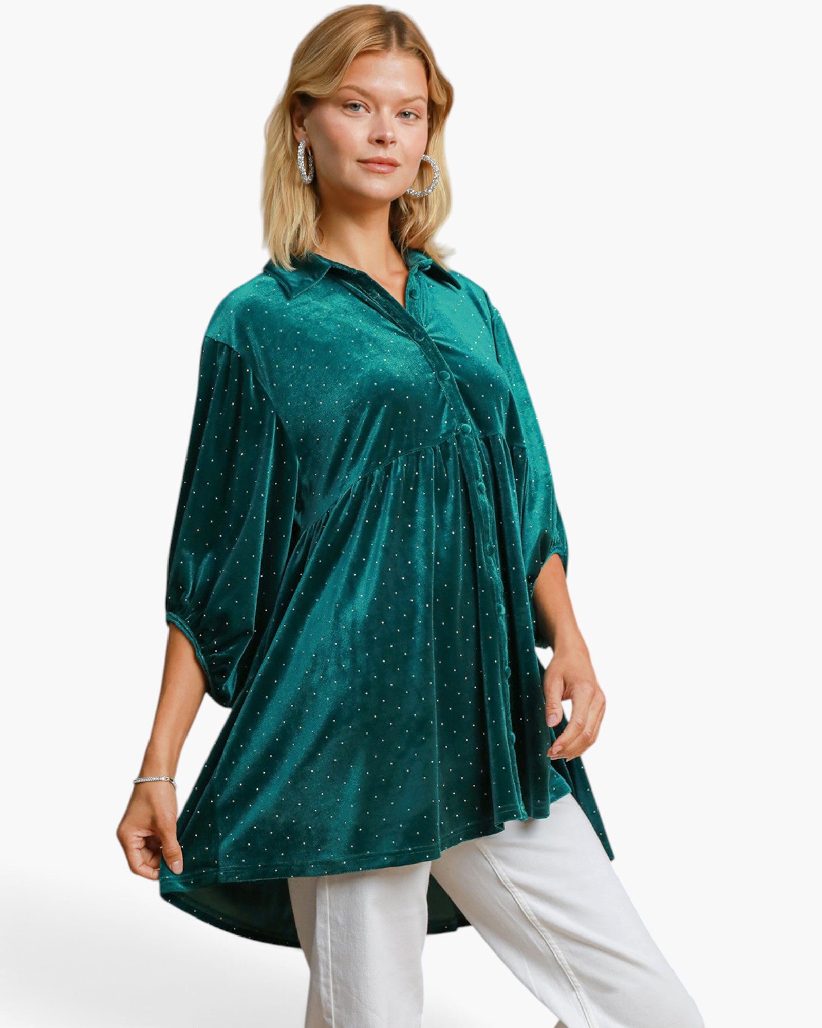 A woman modeling a dark green velvet tunic top with small gold polka dots, paired with white pants, against a plain white studio background.