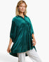 A woman modeling a dark green velvet tunic top with small gold polka dots, paired with white pants, against a plain white studio background.