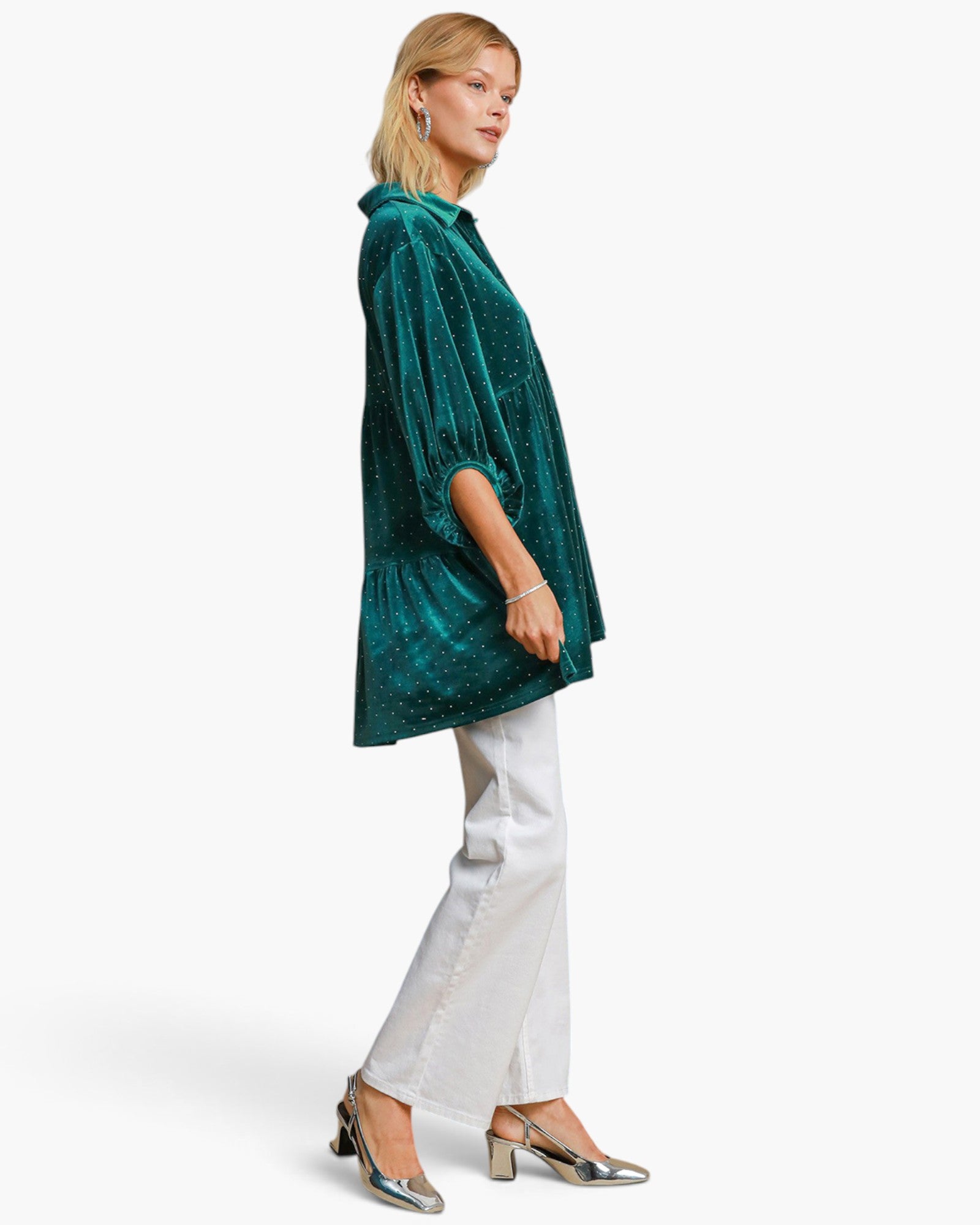 A woman modeling a shimmering emerald green velvet tunic top with elbow-length sleeves, paired with white pants and metallic silver heels, set against a plain white studio background.