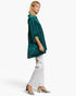 A woman modeling a shimmering emerald green velvet tunic top with elbow-length sleeves, paired with white pants and metallic silver heels, set against a plain white studio background.