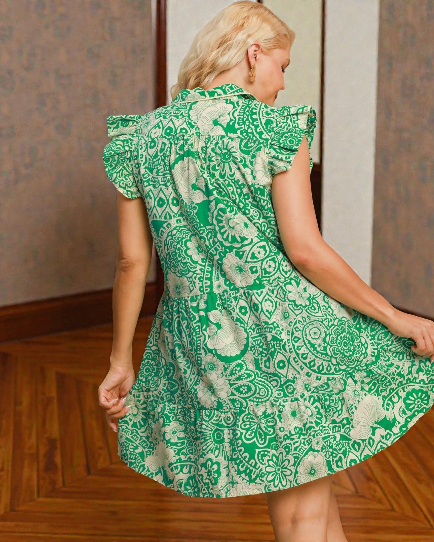 Woman wearing a green patterned dress standing in a room with wooden floor and wallpaper.