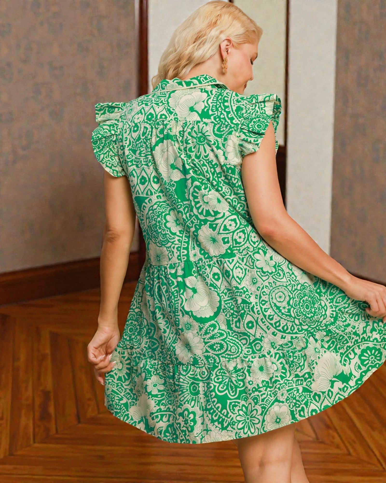 Woman wearing a green patterned dress standing in a room with wooden floor and wallpaper.
