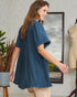 Woman wearing a blue dress in an indoor setting with a plant and painting in the background