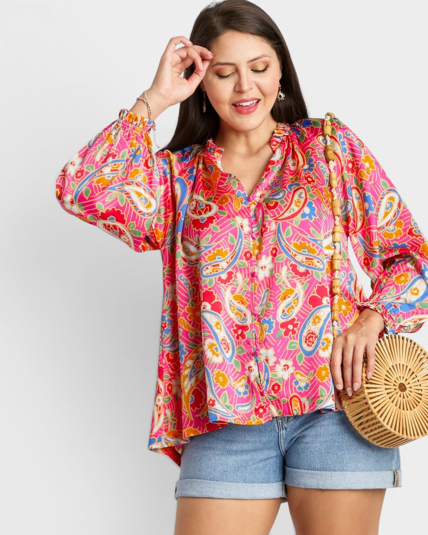 Woman wearing a colorful paisley blouse and denim shorts, holding a woven bag against a white background.