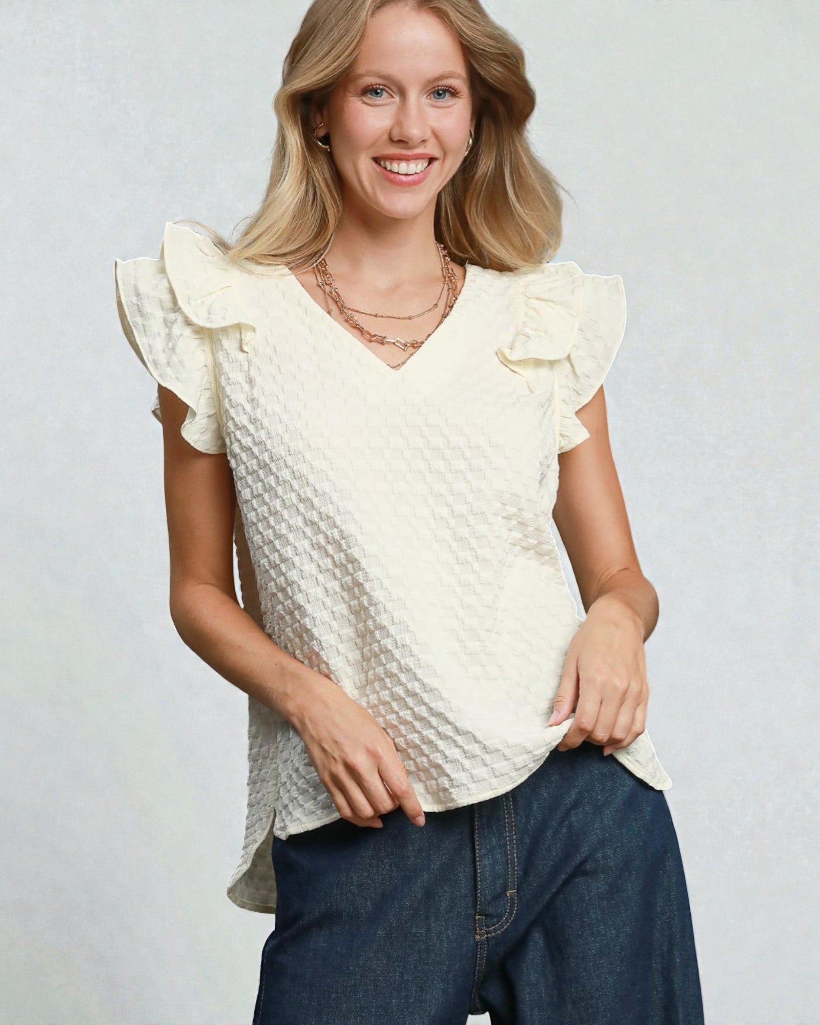 Woman wearing a white textured top with ruffled sleeves and blue jeans on a light gray background