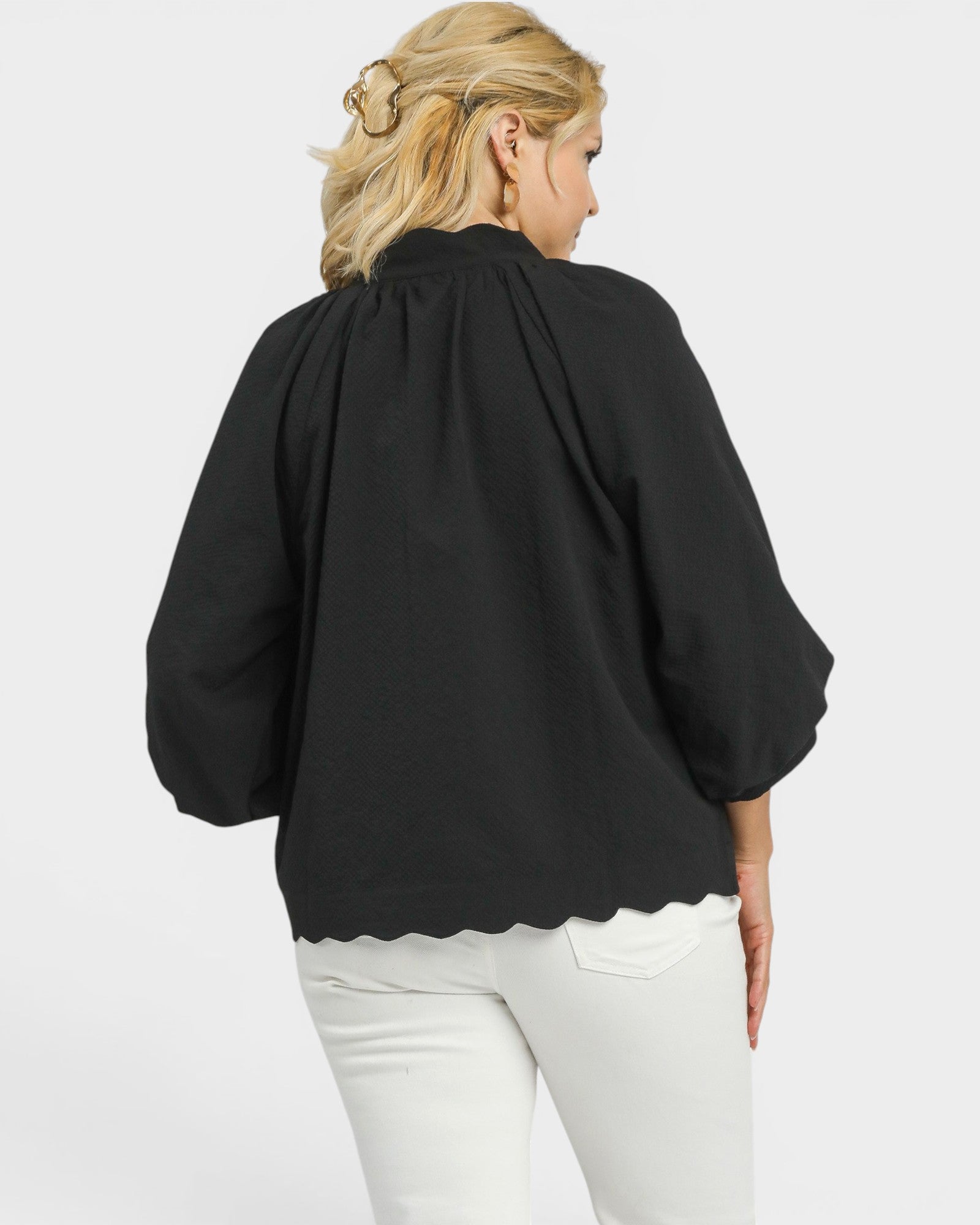 Person wearing a black blouse with scalloped hem and white pants on a light gray background