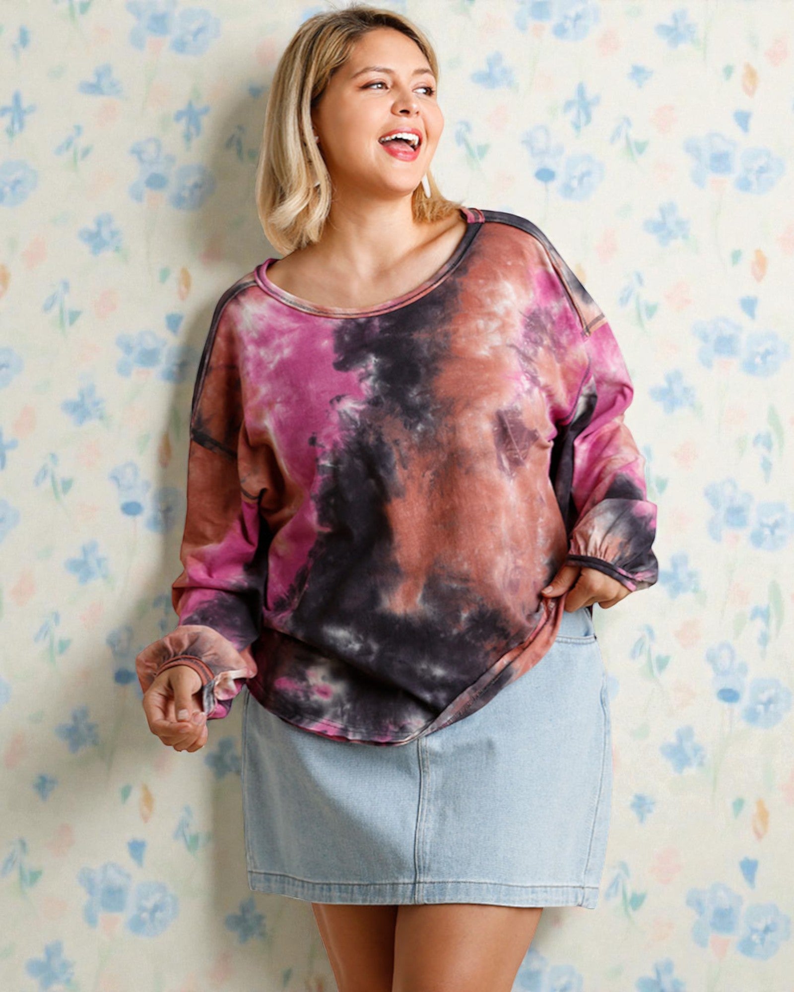 Woman wearing a tie-dye shirt and denim skirt against a floral-patterned wall.