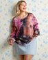 Woman wearing a tie-dye shirt and denim skirt against a floral-patterned wall.