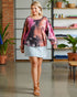 Woman wearing a tie-dye shirt and denim skirt in a room with plants and shelves.
