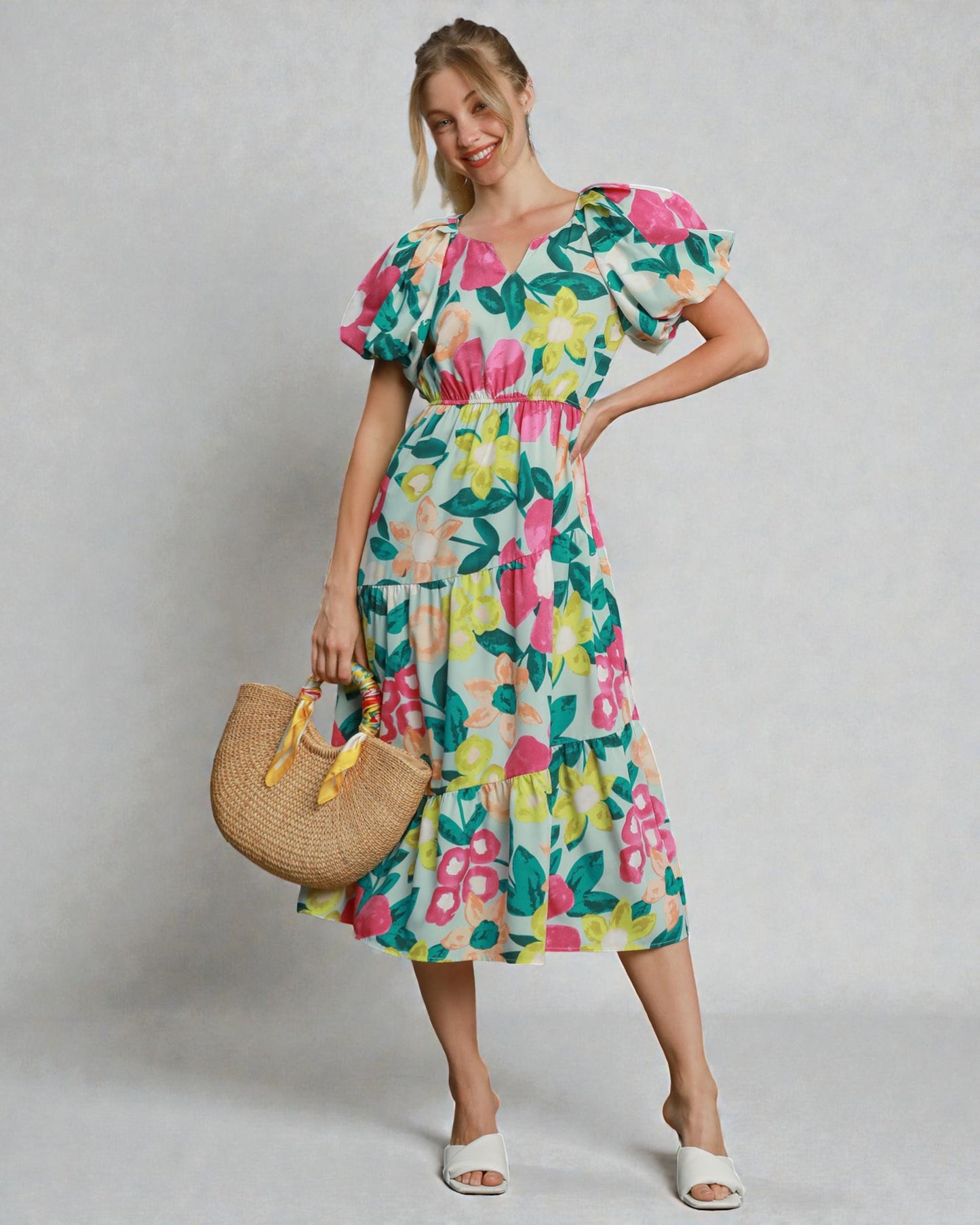 Woman wearing a colorful floral dress holding a straw bag against a plain background