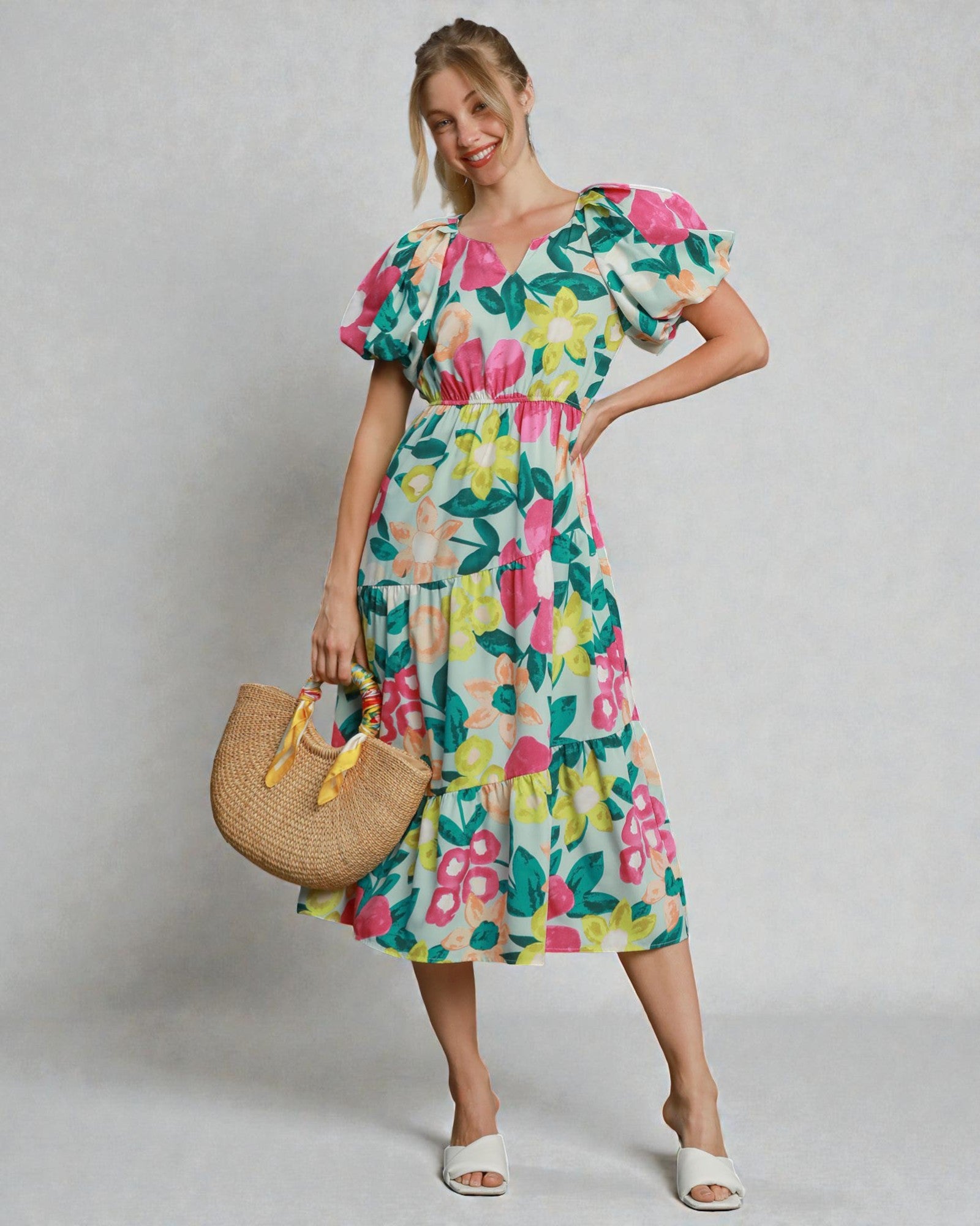 Woman wearing a colorful floral dress holding a straw bag against a plain background