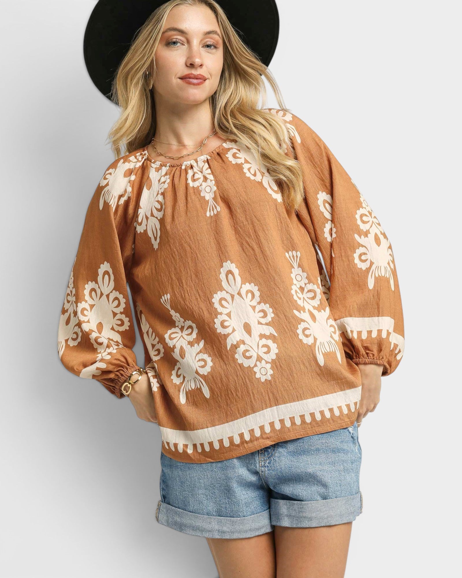 Woman wearing a brown blouse with white patterns and denim shorts on a plain background