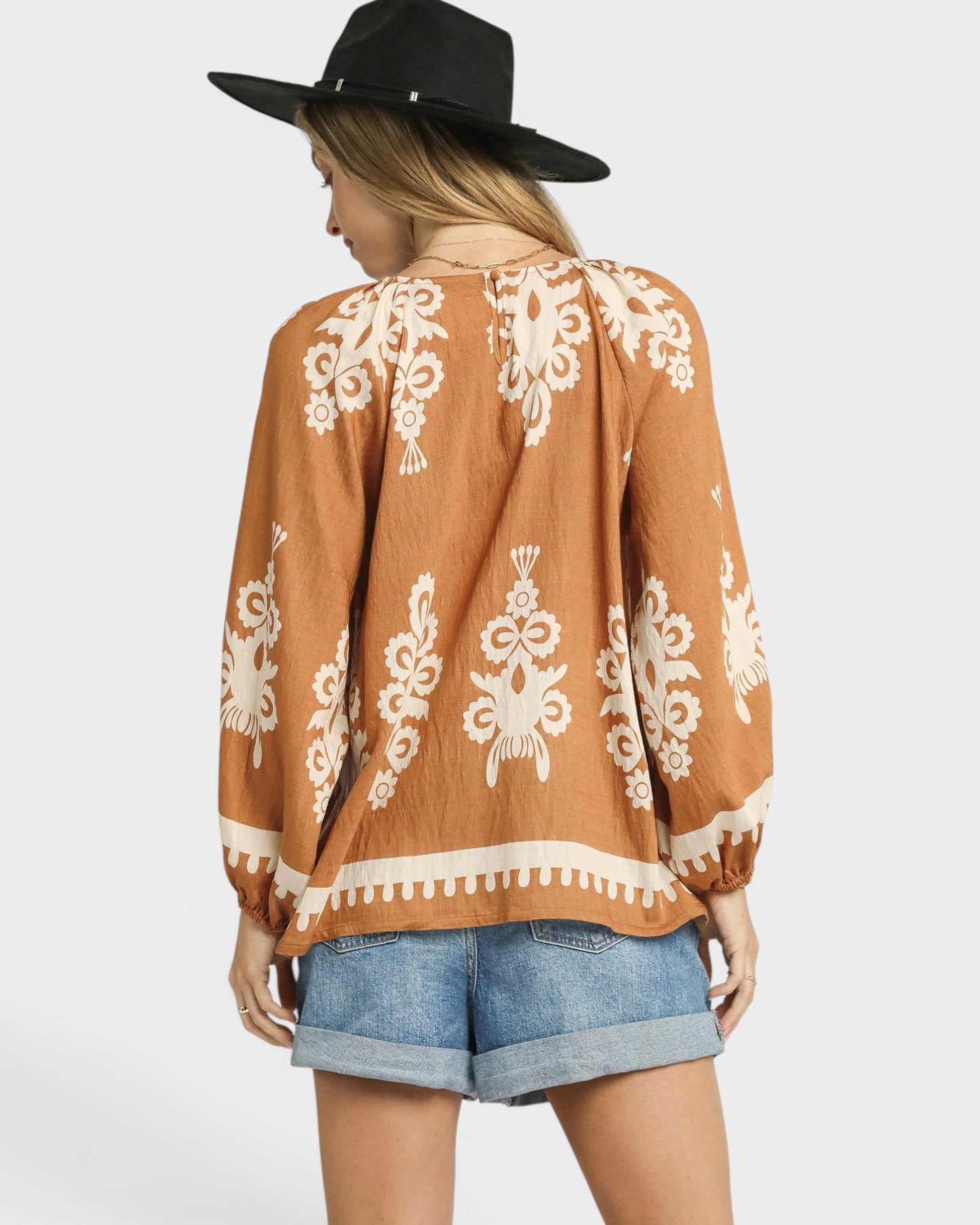 Woman wearing a brown patterned top and denim shorts with a black hat on a white background