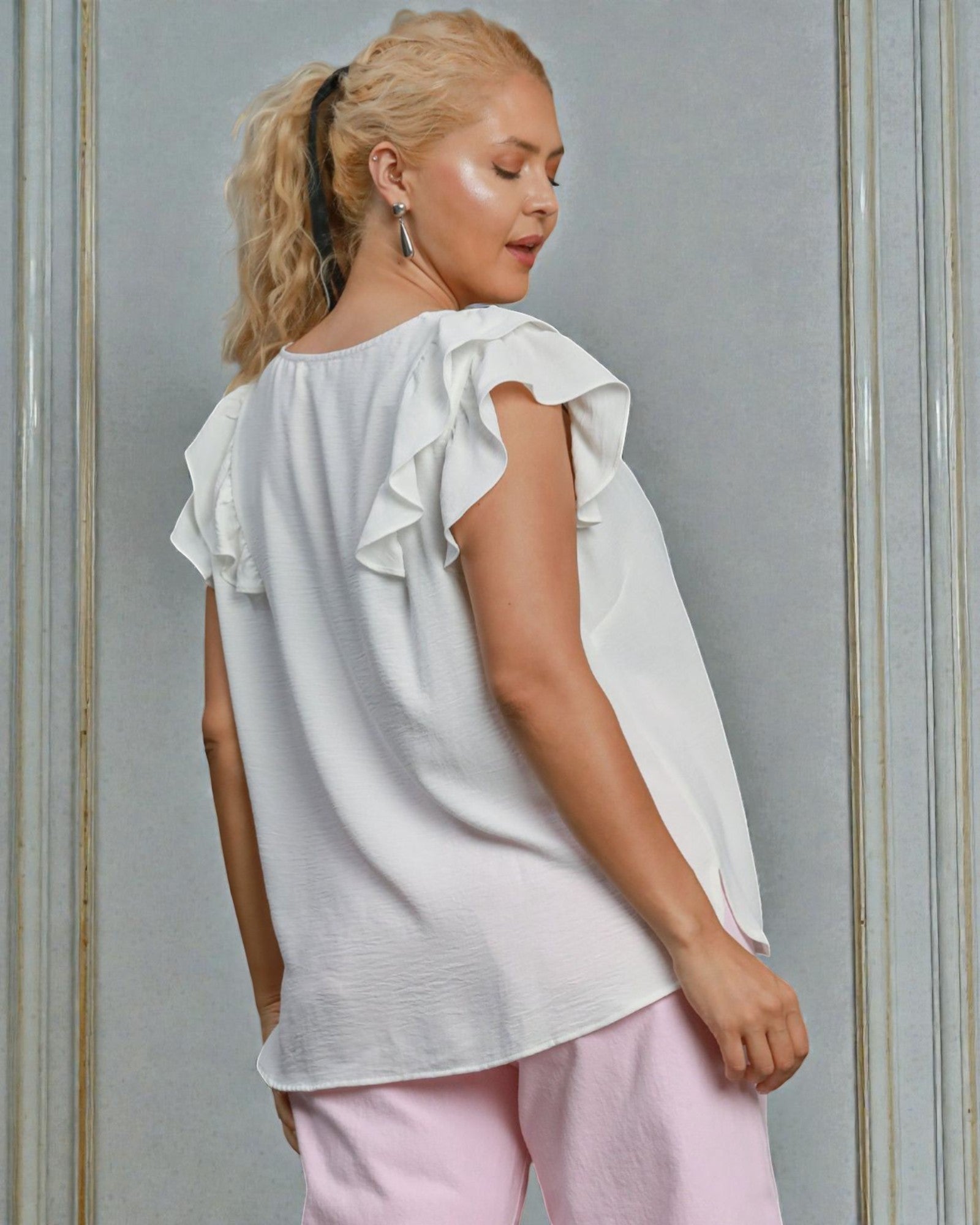Woman wearing a white blouse with ruffled sleeves and pink pants against a gray wall.