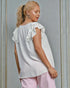 Woman wearing a white blouse with ruffled sleeves and pink pants against a gray wall.