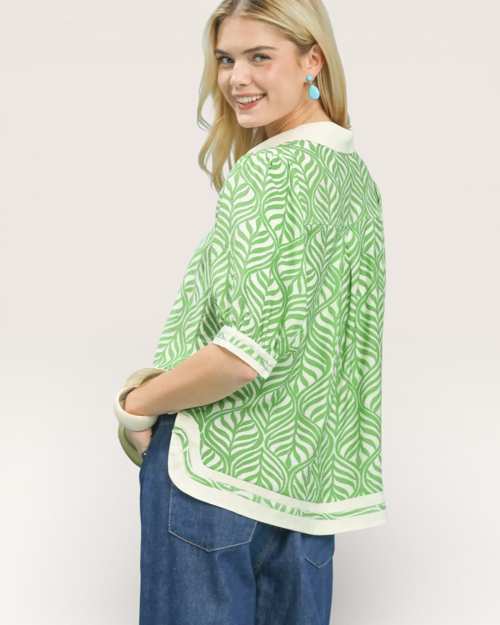 Woman wearing a green and white patterned top with blue jeans on a plain background
