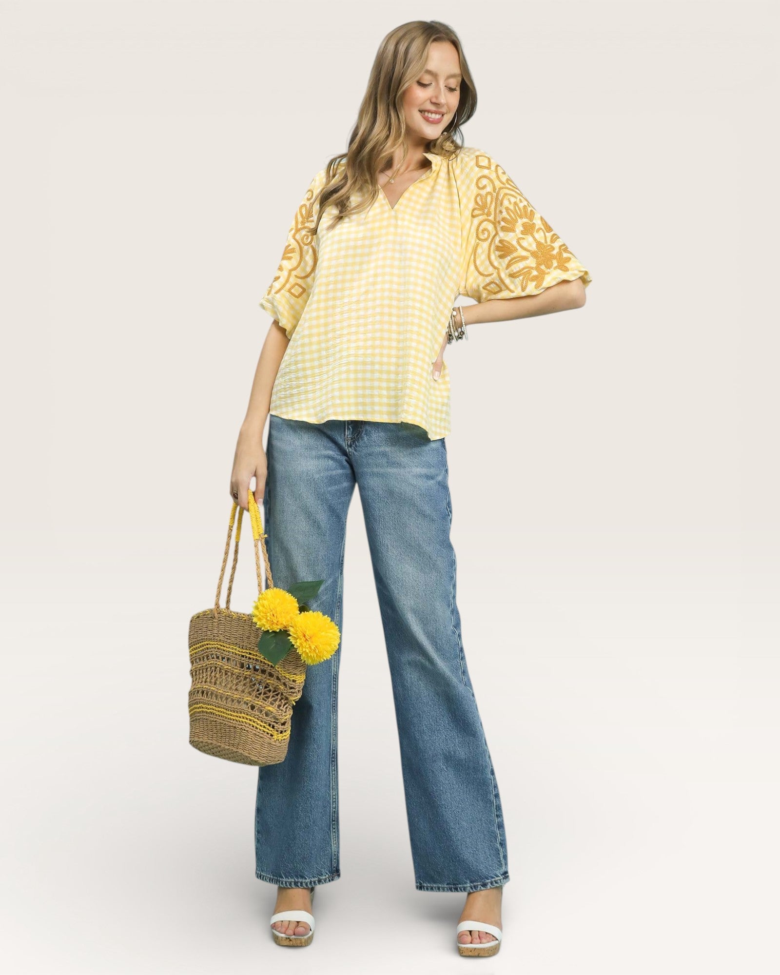 Woman wearing a yellow top with floral patterns and blue jeans, holding a woven bag with flowers.