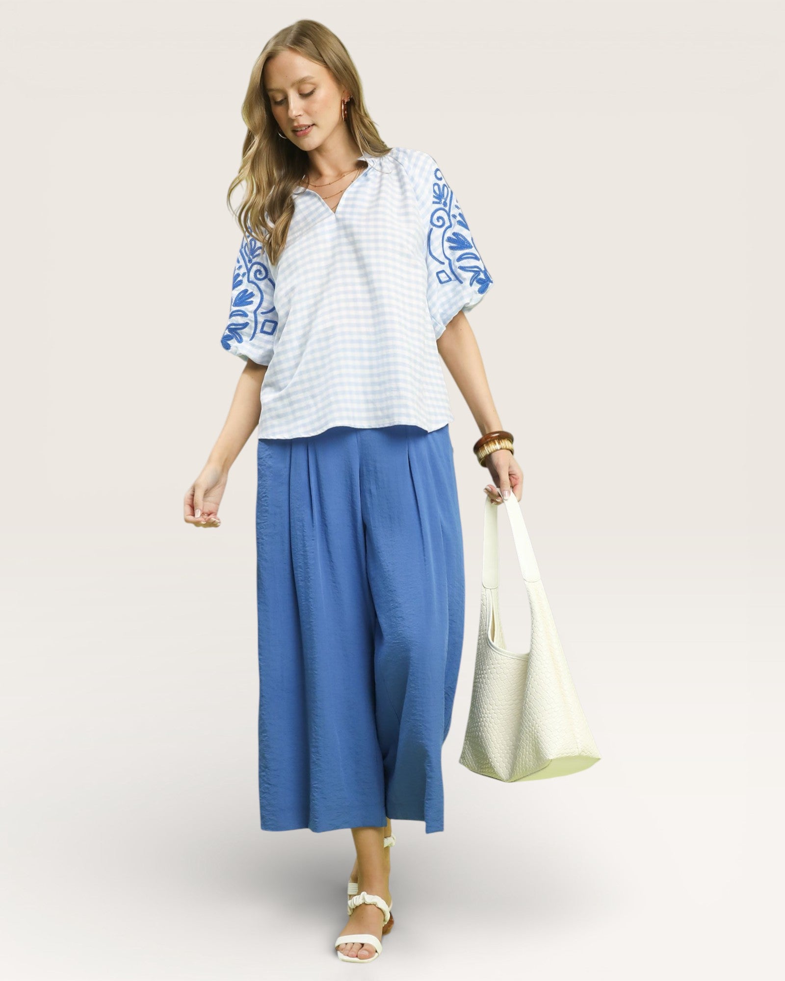 Woman wearing a white blouse with blue embroidery and blue pants, holding a white bag on a light background