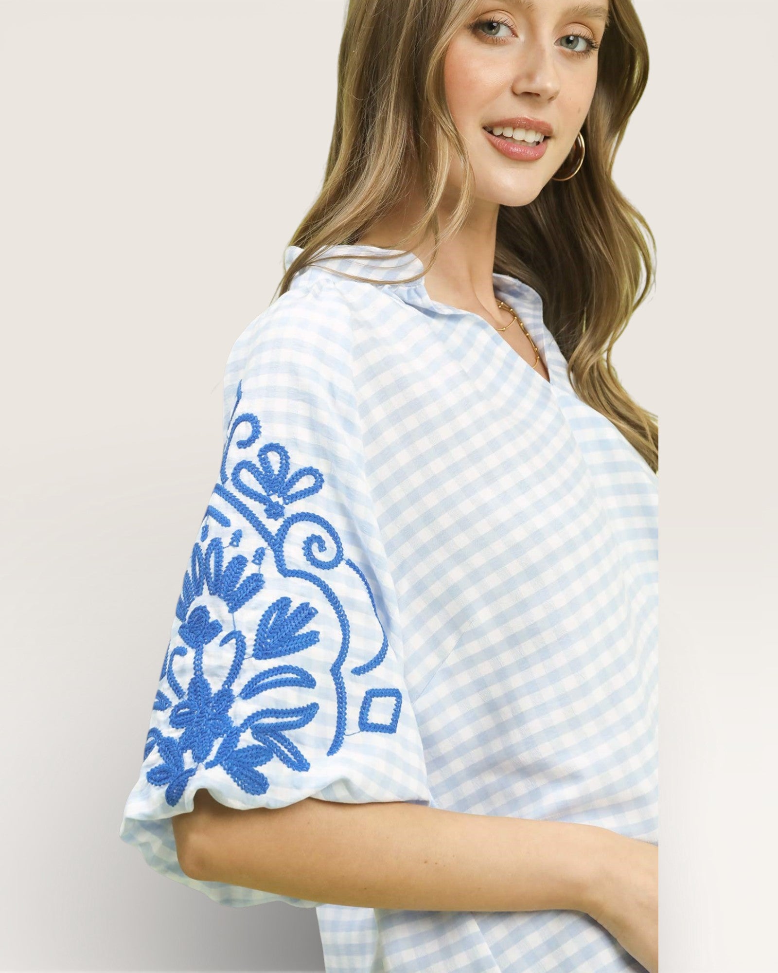 Woman wearing a light blue and white checkered top with floral embroidery on a plain background