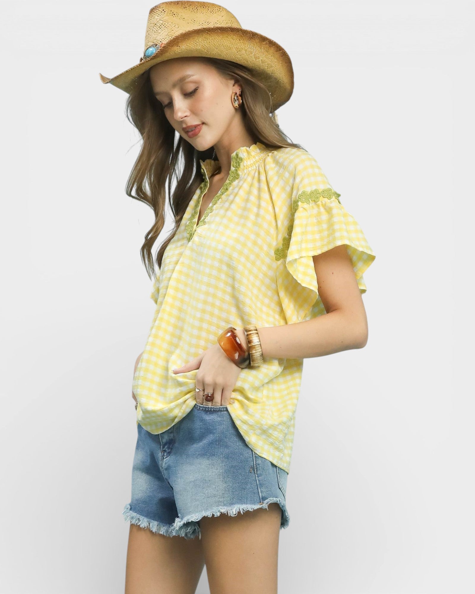 Woman wearing a yellow checkered shirt, denim shorts, and a straw hat on a light gray background