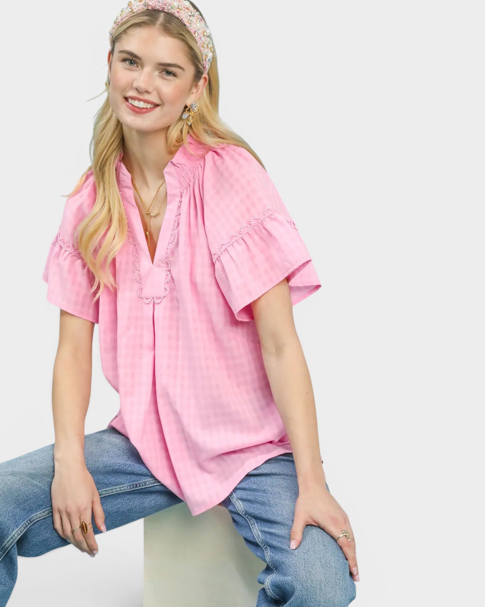 Woman wearing a pink blouse and jeans sitting on a white stool against a white background