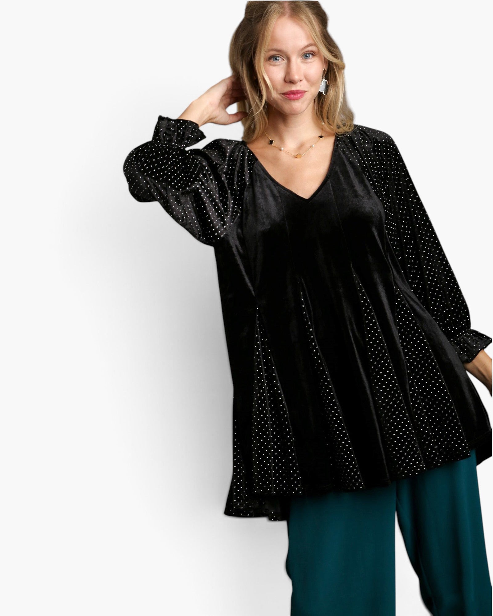 A woman modeling a black velvet long-sleeve top with dotted sheer overlay sleeves and side panels, paired with teal pants, against a plain white studio background.