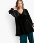 A woman modeling a black velvet long-sleeve top with dotted sheer overlay sleeves and side panels, paired with teal pants, against a plain white studio background.
