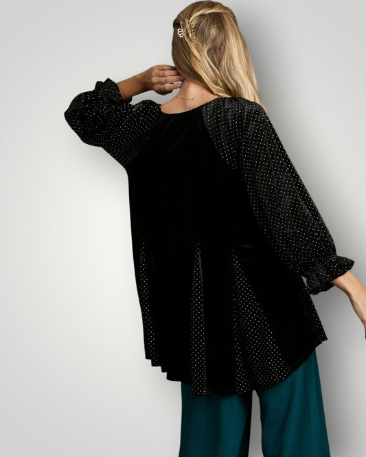 Woman wearing a black polka dot blouse with long sleeves and teal pants on a plain background