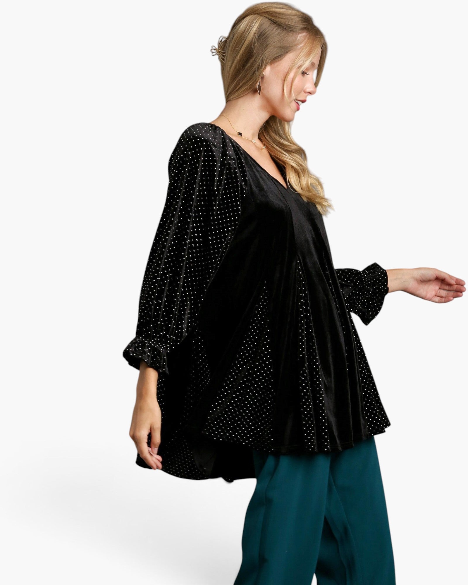 A woman wearing a black velvet long-sleeve top with white polka-dot pattern on the sleeves and hem, paired with teal pants, photographed against a clean white studio background.