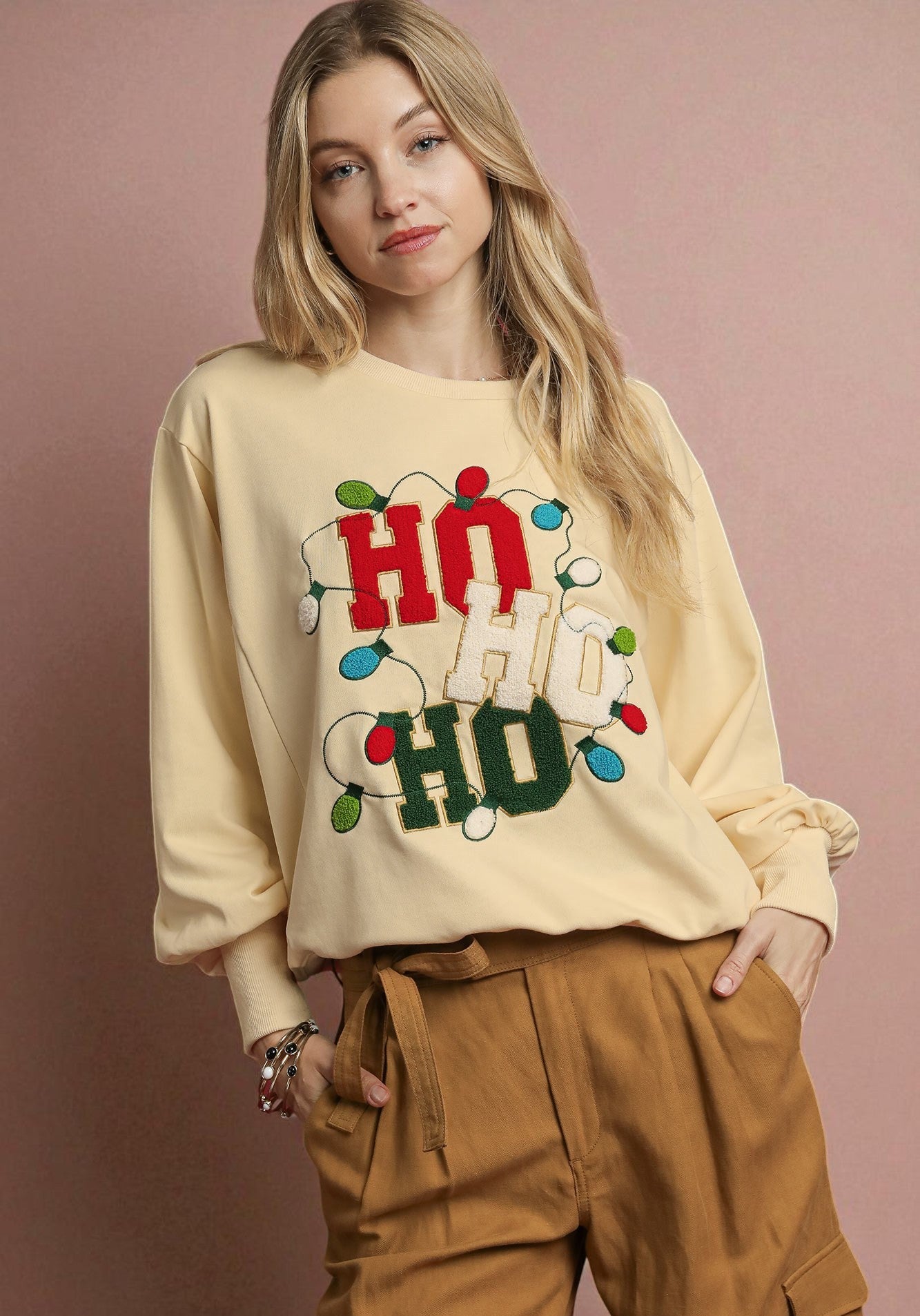 Woman wearing a beige sweatshirt with 'Ho Ho Ho' text and lights on a pink background