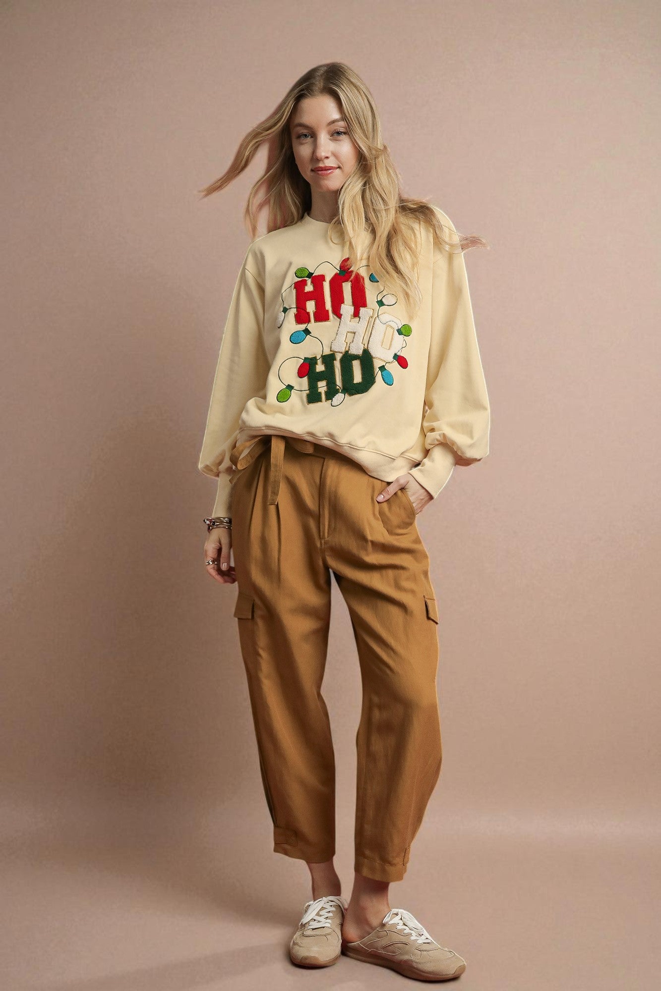 Woman wearing a beige sweater with colorful text and brown pants on a beige background