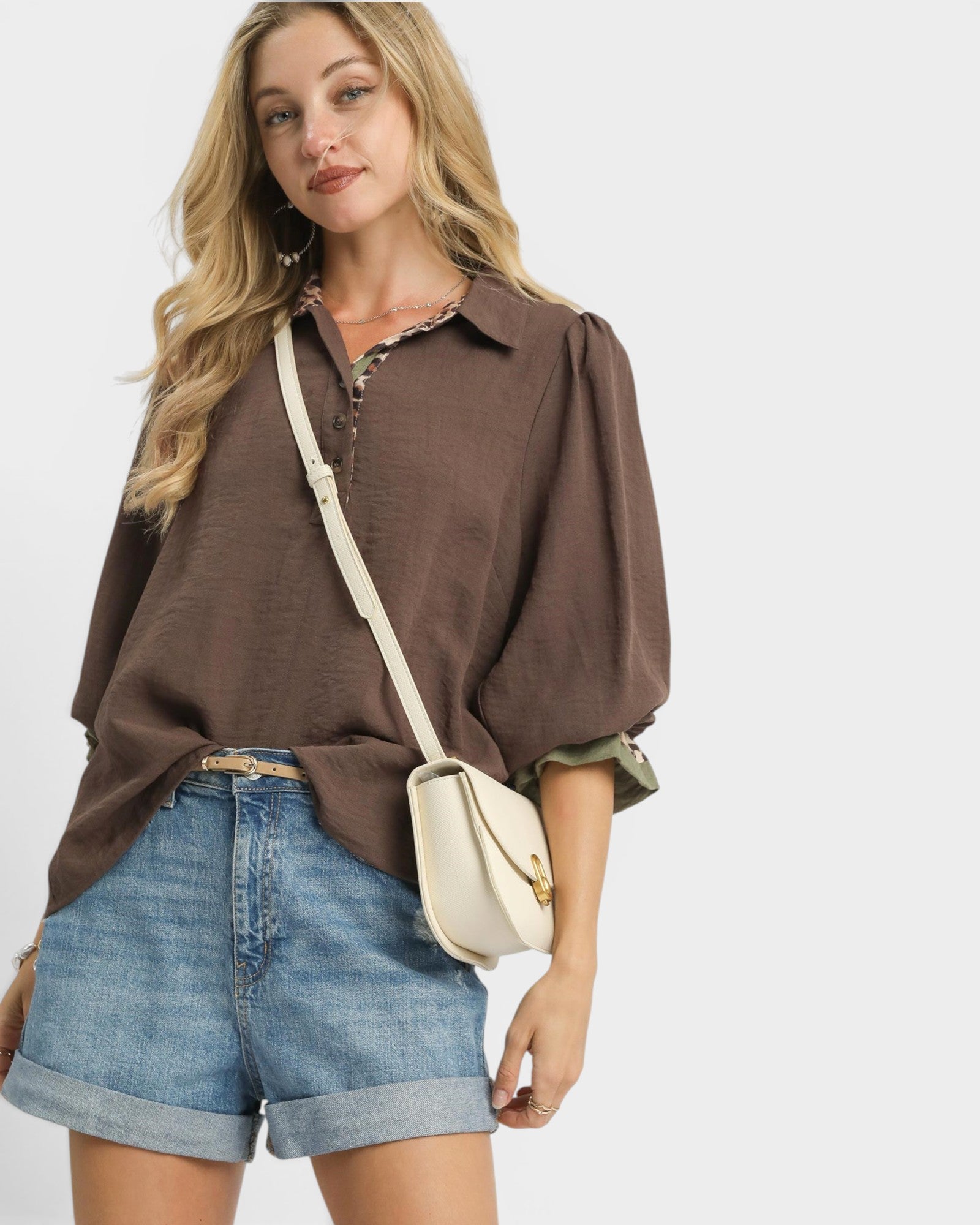 Woman wearing a brown blouse and denim shorts with a beige crossbody bag on a white background