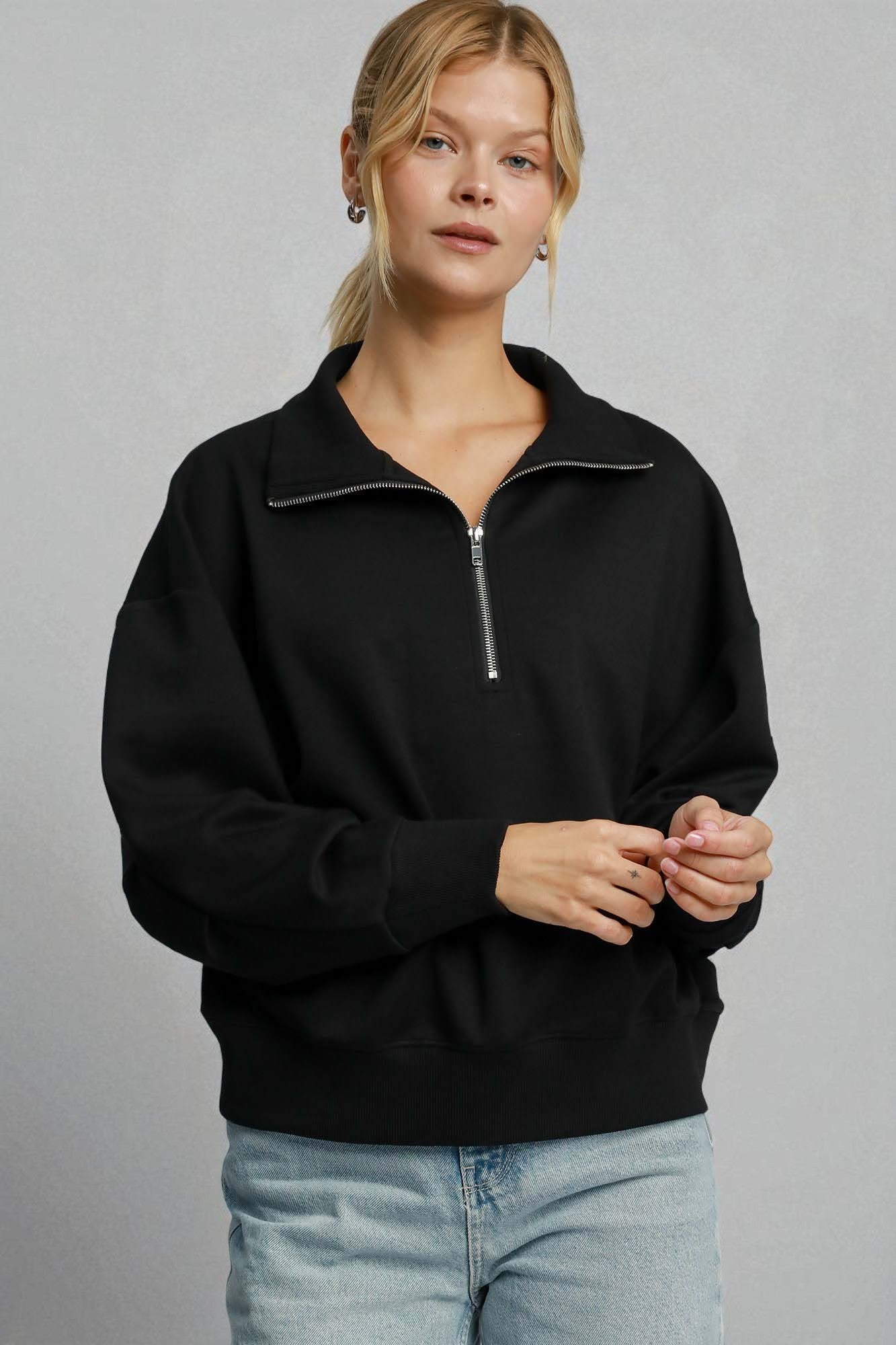 Woman wearing a black quarter-zip sweatshirt and light blue jeans against a gray background