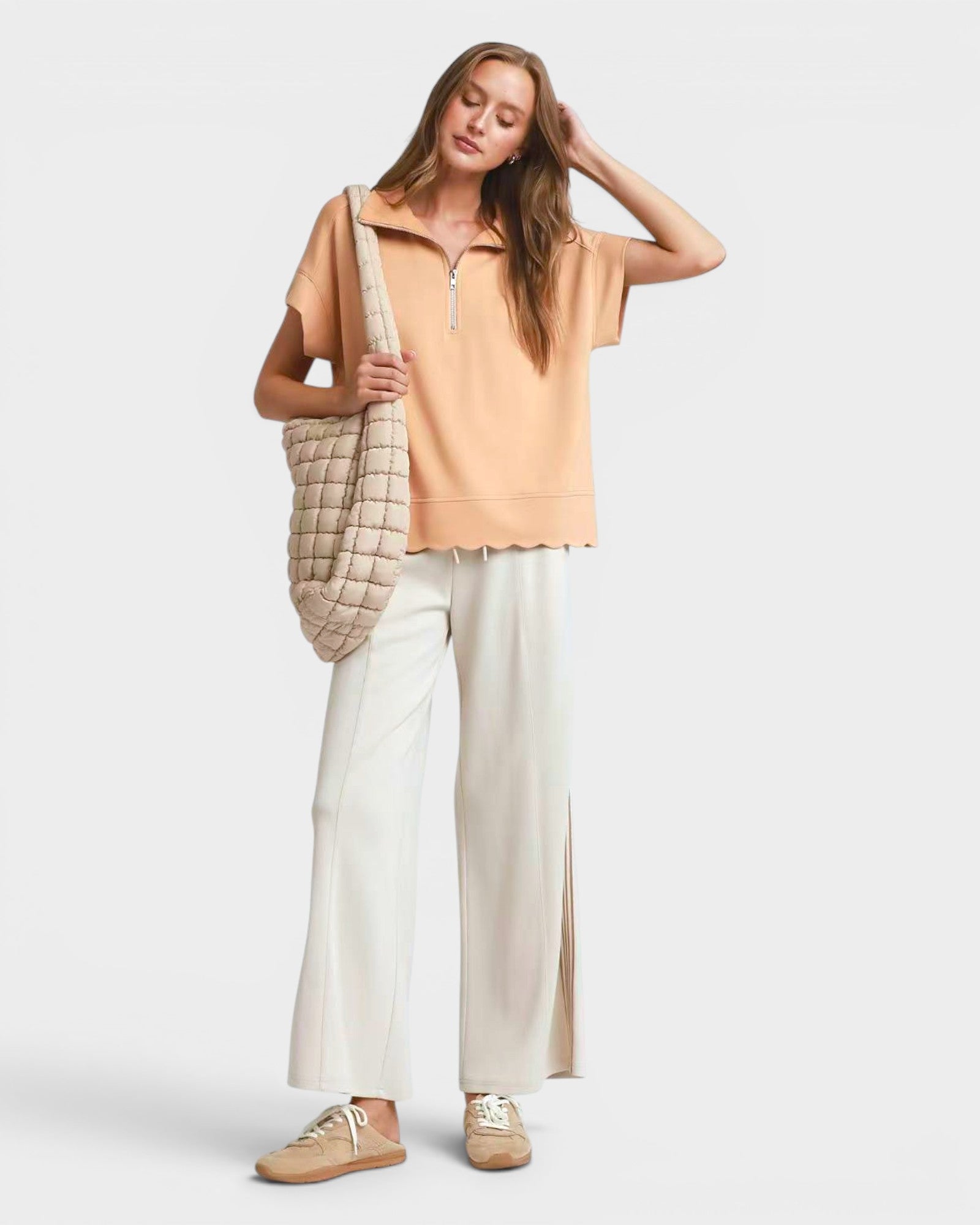 Woman wearing a beige top and white pants, holding a checkered bag on a light gray background