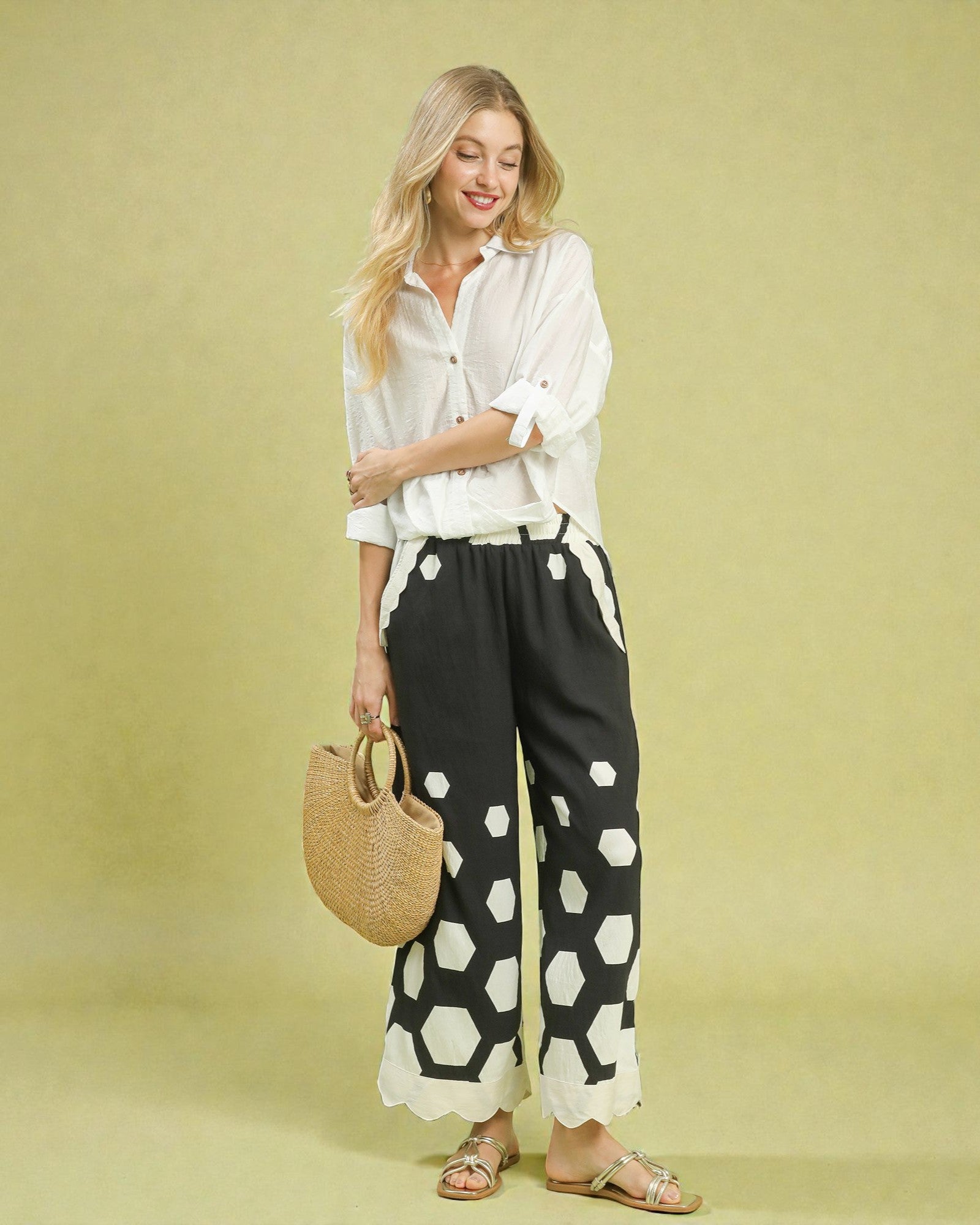 Woman wearing a white shirt and black pants with white patterns, holding a woven bag on a yellow background