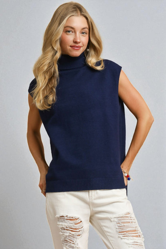Woman wearing a navy sleeveless sweater and white pants against a gray background