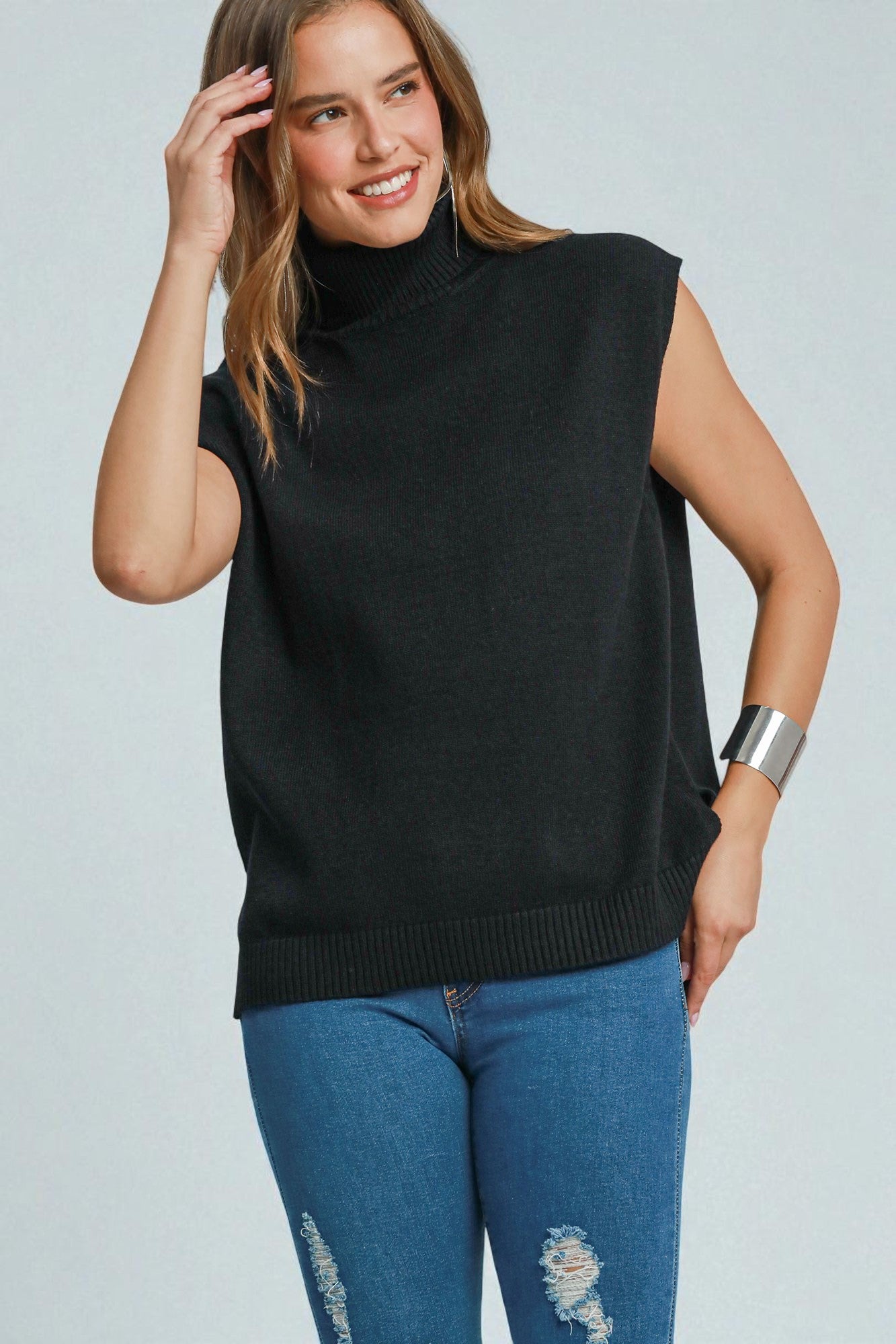 Woman wearing a black sleeveless sweater and blue jeans against a light gray background