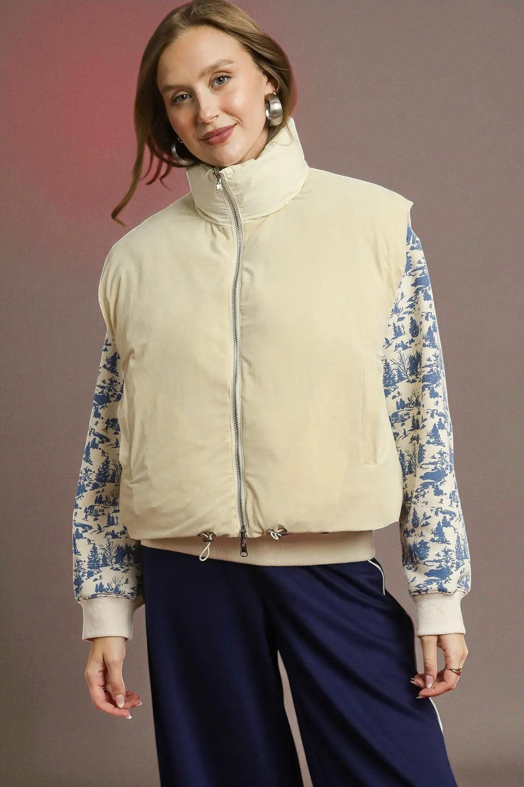 Woman wearing a beige puffer vest over a floral-patterned shirt with navy pants against a gradient background.