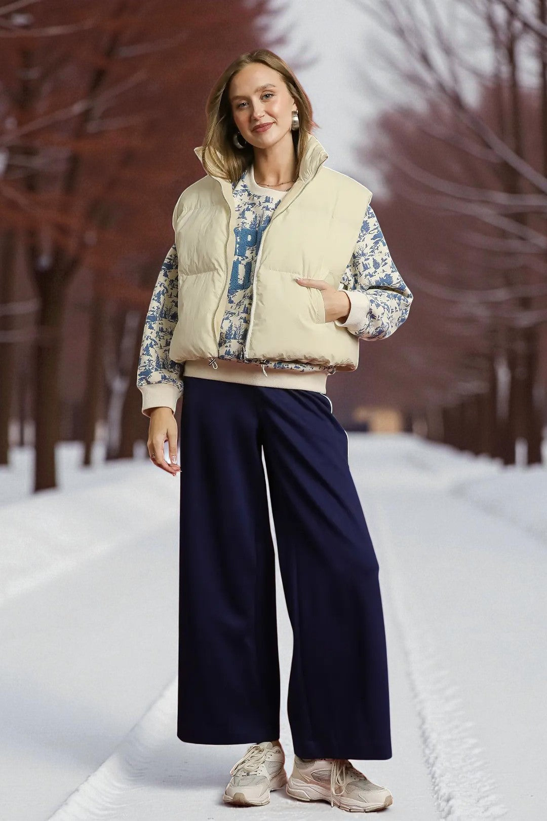 Woman wearing a light-colored vest, patterned jacket, and dark pants in a snowy landscape.