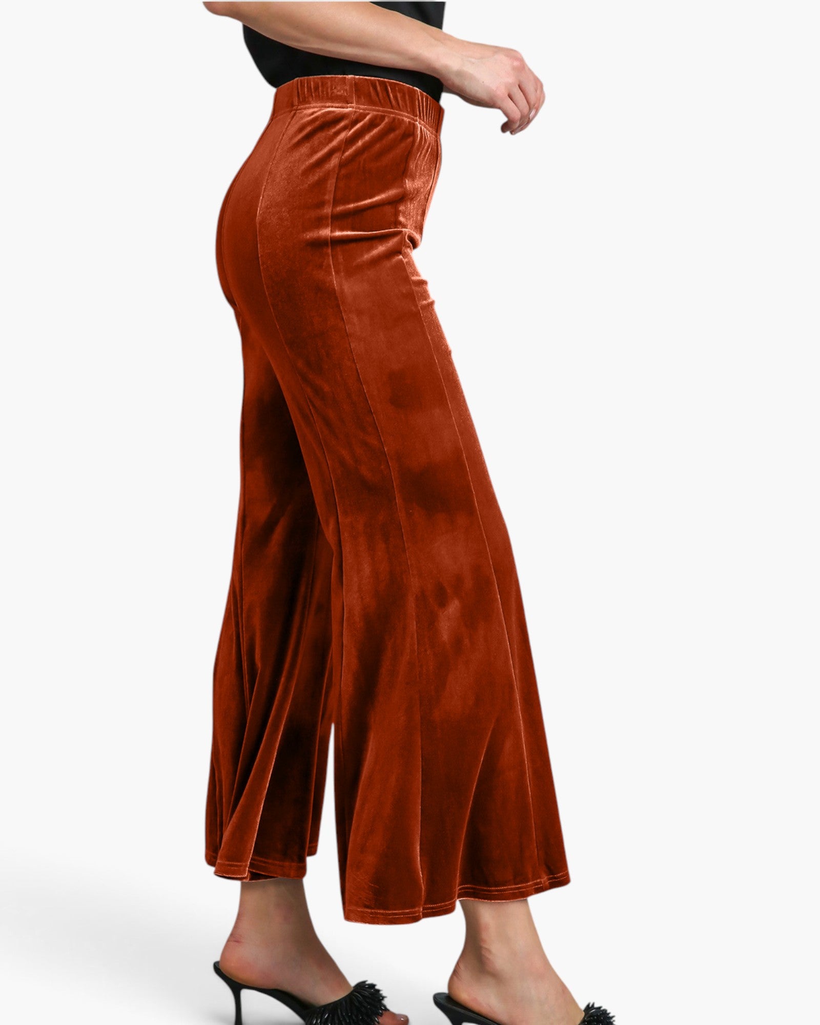 A person wearing rust-orange velvet wide-leg cropped pants with a black top and black heeled mules, shown from the waist down against a light gray studio background.