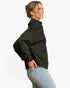 A woman shown from the side profile wearing a black oversized pullover and light blue jeans against a plain white studio background.