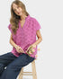 Woman wearing a pink embroidered blouse sitting on a stool with flowers, against a plain background
