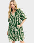 A woman modeling a green and cream tropical leaf print short-sleeve dress against a light gray background.