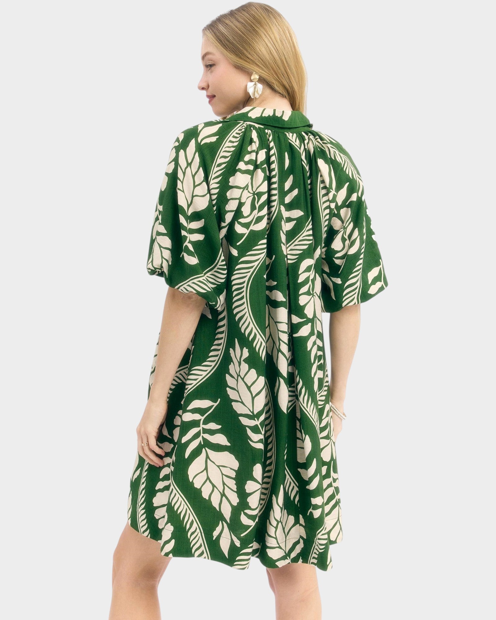 A woman wearing a green and cream tropical leaf print dress, shown from the back against a light gray studio background.