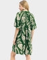 A woman wearing a green and cream tropical leaf print dress, shown from the back against a light gray studio background.