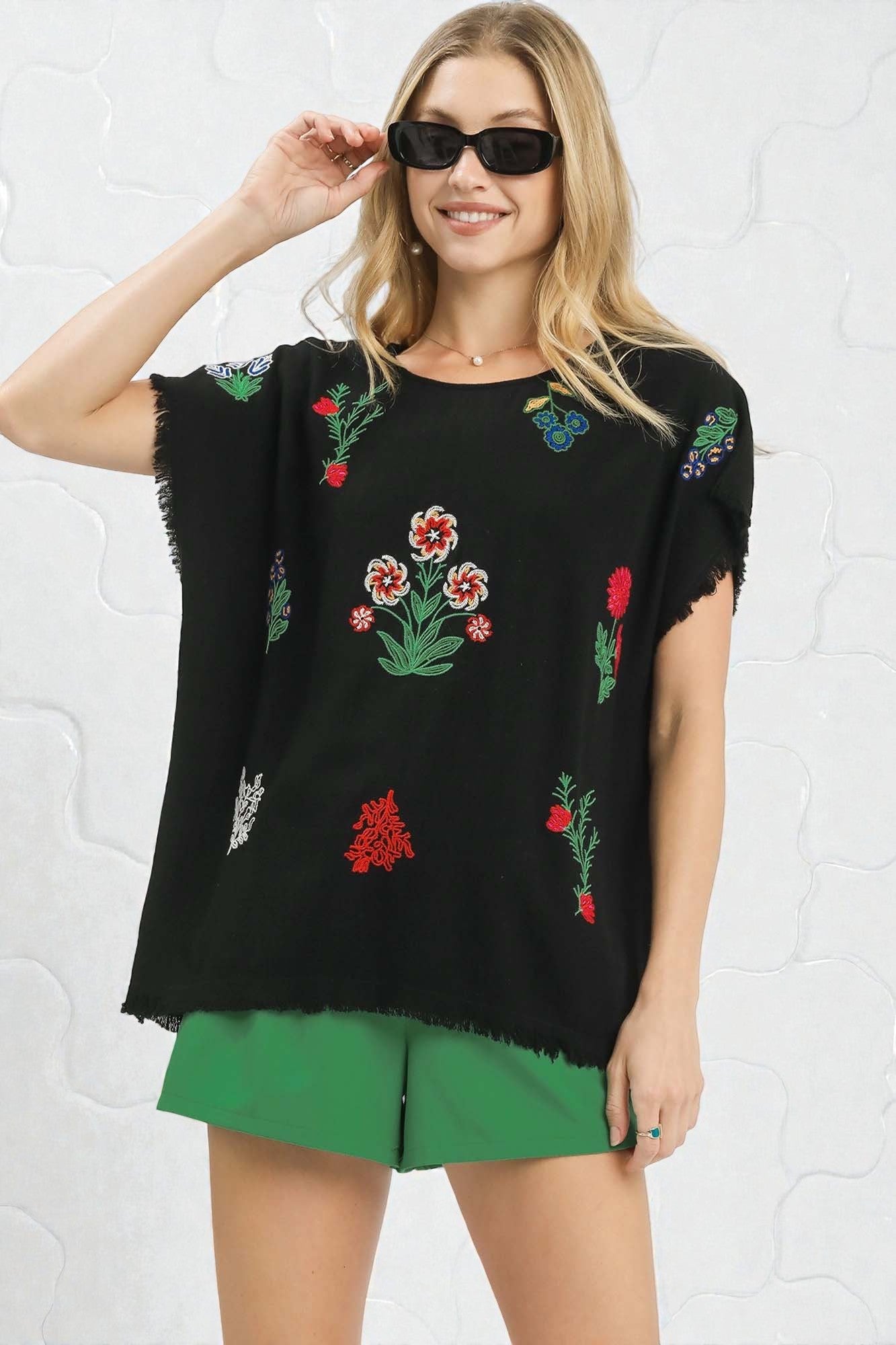 Woman wearing a black top with colorful floral embroidery and green shorts against a white brick wall.